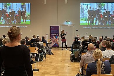 Ein großer Raum, in dem viele Menschen sitzen, auf der Bühne steht eine Frau. An der Wand wird ein Bild vom A capella Ensemble Thonkunst gezeigt