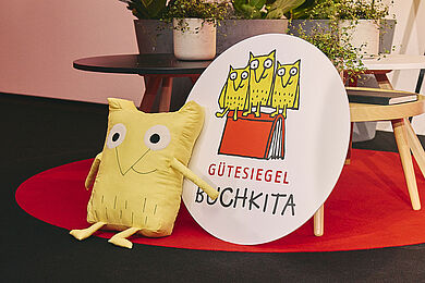 Links Eulen-Kuscheltier und daneben ein großes rundes Holzschild mit Logo und Aufschrift "Gütesiegel Buchkita"