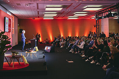 Blick in den Raum bei der Preisverleihung, gedimmtes Licht, links ein Moderator, rechts Blick auf das Publikum