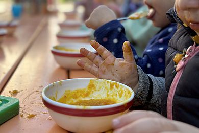 Einige Kinder sitzen am Tisch und lassen sich die Suppe schmecken, die in kleinen Schüssel angereicht wird. An den kleinen Hände und Mündern zeigen sich auch die Spuren der Suppe.