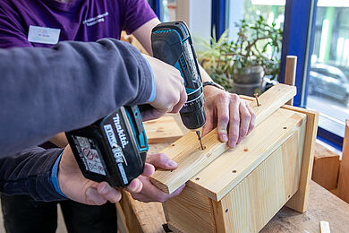 Eine Person schraubt mit einem Akkuschrauber eine Latte an die Rückseite eines Vogelhäuschens aus Holz, das auf einer Werkbank liegt. Eine weitere Person hält die Latte fest. Man sieht nur die Unterarme der Personen.