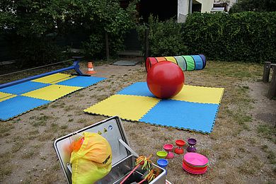 Im Garten liegen auf bunten Spielmatten ein großer roter Hüpfball, Flowersticks zum Jonglieren und Diabolos für die Kinder bereit.