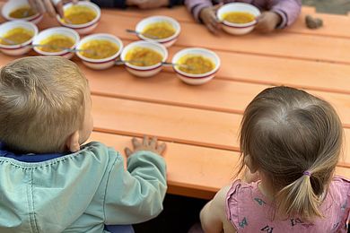 Ein Junge und ein Mädchen sitzen mit dem Rücken zur Kamera gewandt nebeneinander am Tisch. Der Junge trägt ein hellblaues Jäckchen mit Kapuze. Das Mädchen hat ein rosa Oberteil mit Schmetterlingen an. Gegenüber sitzen andere Kinder und essen die Suppe aus den Schüssel. 