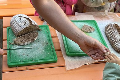 Die Hand eines Erwachsenen reicht ein Stück Brot an eine Kinderhand. Im Hintergrund liegen zwei Brote auf Brettchen. Eines wurde bereits angeschnitten, das andere ist unberührt.