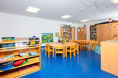 Leseecke im Gruppenraum, in der MItte des Raumes stehen 2 Tische mit Stühlen, links ein Regal mit Büchern
