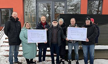 Mehrere Personen stehen vor dem roten Gebäude der BBW-Kita "Gustav-Freytag-Straße 31". Die Personen halten zwei Spendenschecks in den Händen. Sie sehen fröhlich aus und freuen sich über die Übergabe. Die Menschen sind warm angezogen und auf dem Boden liegt noch etwas Schnee.