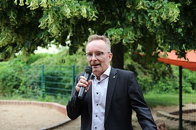 Der Markkleeberger Bürgermeister Olaf Schlegel findet an diesem besonderen Tag ebenfalls anerkennende Worte für das Kita-Team. An seinem schwarzen Sakko trägt er eine Anstecknadel mit dem Wappen der Stadt Markkleeberg und eine Brille, die über den Augen schwarz gerahmt ist.