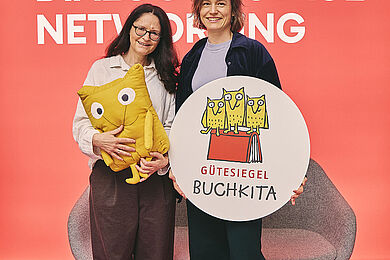 Zwei Frauen vor einer orangenen Wand. Die linke Frau hält ein Kuscheltier in Form einer Eule, die rechte ein rundes Siegel aus Holz mit der Aufschrift Gütesiegel Buchkita