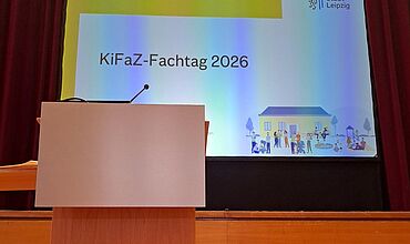 Auf einem großen Bildschirm auf einer Bühne steht „KiFaZ-Fachtag 2026“, daneben sind das Logo der Stadt Leipzig und eine Illustration mit einem Haus und vielen Meschen drum herum. Vor der Bühne steht ein Rednerpult.
