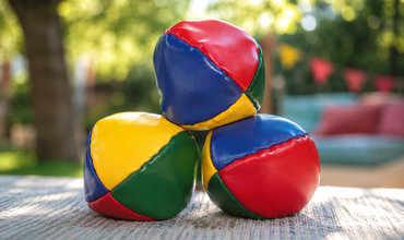 3 bunte Jonglierbälle liegen im Vordergrund auf einem Tisch. Im Hintergrund Bäume und eine bunte Girlande, ein blaues Sofa mit roten Kissen.
