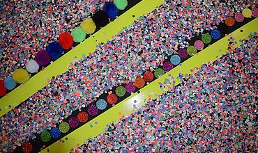 Auf einer Leinwand sind durch Klebeband diagonale Bereiche abgetrennt, die mit Konfetti, Glitzerpunkten und Stoffbommeln beklebt sind