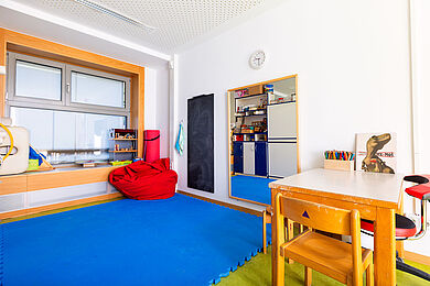 "Therapieraum" mit weicher Bodenmatte, rotem Sitzsack. Rechts steht ein Tisch auf dem Buntstifte in einem Stifthalter stehen.