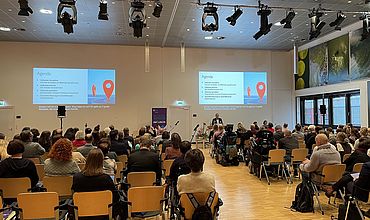 In einem großen Saal sind viele sitzende Menschen zu sehen, auf der Bühne hält Prof. Böhm einen Vortrag bei der Inklusionskonferenz. Die Agenda ist durch einen Beamer an die Wand hinter ihm angezeigt.