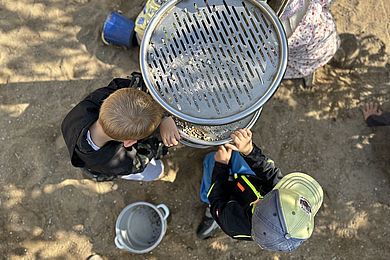 Zwei Jungs spielen im Sand und lassen kleine Steine durch ein großes Sieb aus Metall rieseln. Man sieht sie nur von oben. Einer von ihnen trägt ein Basecap.