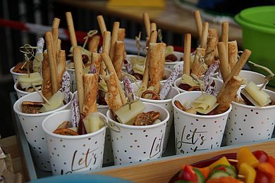 Beim Buffet stehen Pappbecher mit der goldenen Aufschrift Let`s Party. Sie sind gefüllt mit kleinen Salami-Würstchen, Käsehäppchen und Knusperstangen.