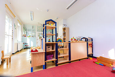 Gruppenraum im Krippenbereich: Im Vordergrund ein roter Teppich. In der Mitte ein Rauteiler aus hellen Holzelementen mit Spielzeug und Spielmaterial, helles, freundliches Licht