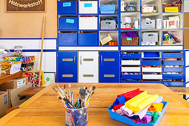 Atelier - im Vordergrund auf dem Tisch Pinsel und Krepppapier, im Hintergrund ein Schrank mit blauen und weißen Schubfächern und Bastelmaterialien. Links oben hängt ein Schild mit der Aufschrift "Kinder Holzwerkstatt"