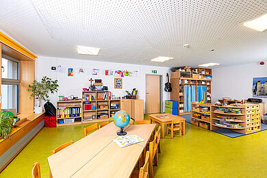 Familiengruppe im 1. OG rechts - Kindergartenraum mit Regalen und im Vordergrund einem Tisch, auf dem ein Globus steht. Es sind Spielmaterialien, Bücher und bunte Bilder erkennbar.