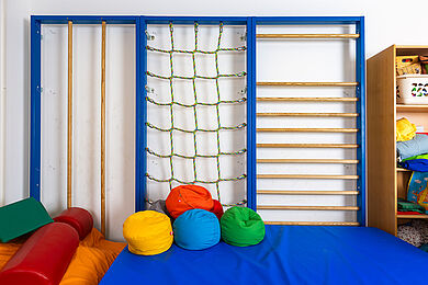 Sprossenwand mit Stangen und Netz im Sportraum, davor eine blaue Matte und mehrere bunte Sitzsäcke/Bälle