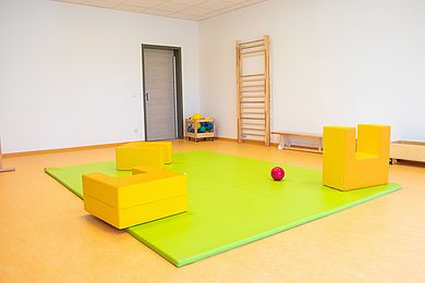 In einem Zimmer liegt eine apfelgrüne Matratze, auf der ein roter Ball und große gelbe Bausteine aus Stoff verteilt sind. Im Hintergrund hängt eine Sprossenwand. 